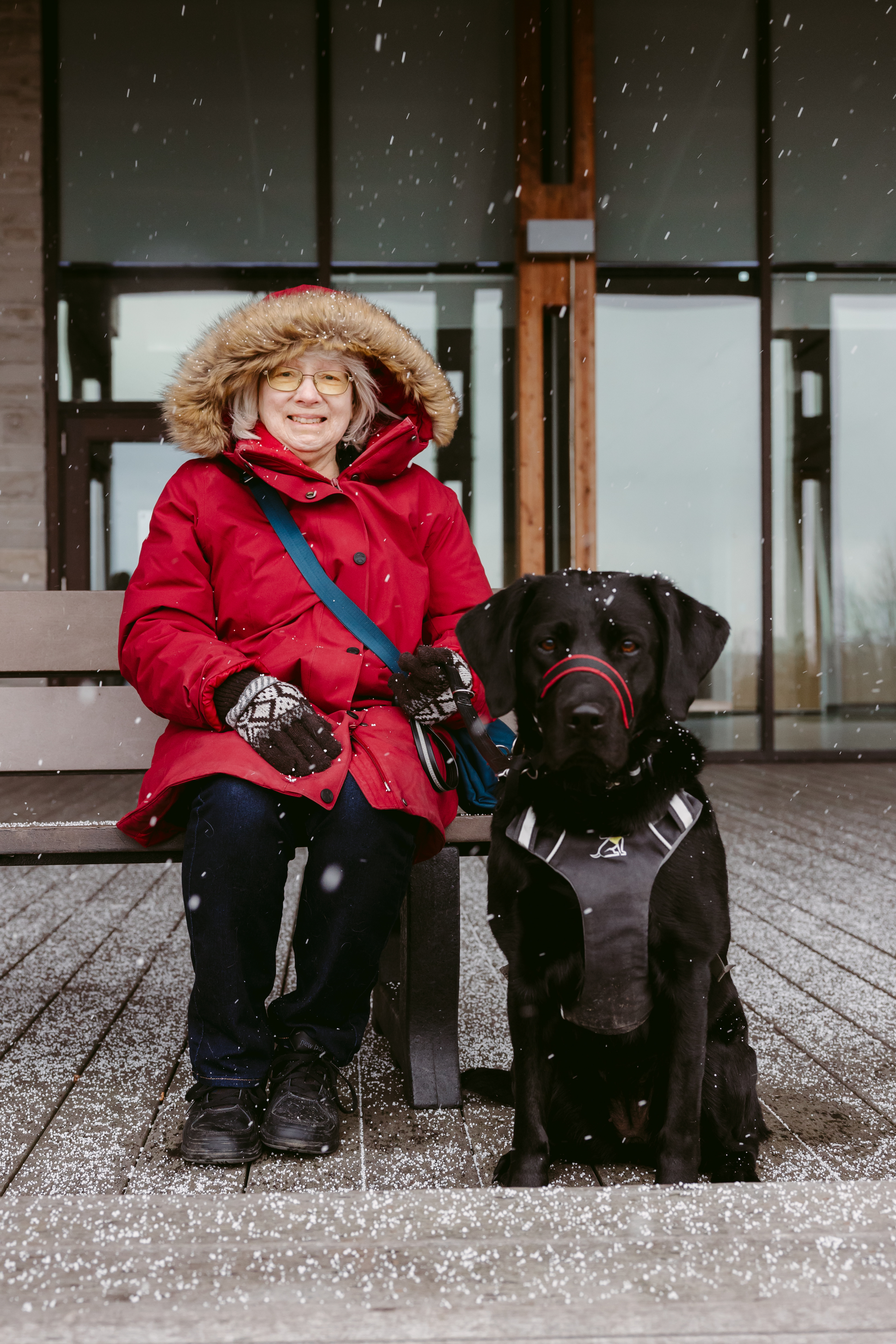 Cheri smiles and sits on a bench, and her guide dog, Sassy, sits on the pavement to her right. Sassy is a black dog in harness. It’s lightly snowing, and Cheri is wearing a red winter jacket and gloves.