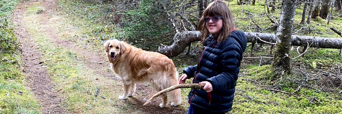 Rhea and her CNIB Buddy Dog Ivy, a two-year-old golden retriever, walking along a trail in the woods. Rhea is holding a stick and they are both looking back at the camera, smiling.