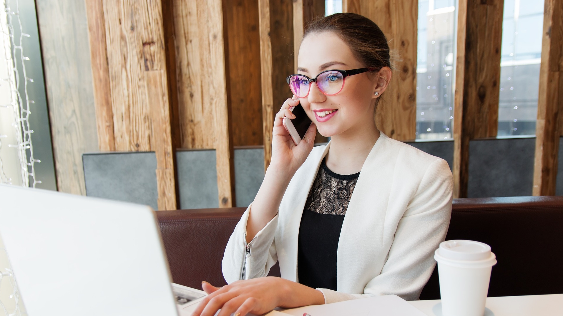 Une jeune femme portant des lunettes parle au téléphone et est sur l'ordi. un café pour emporté est déposé sur la table.