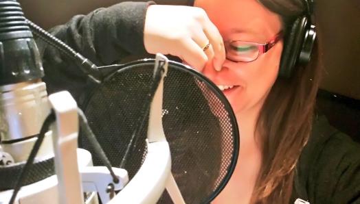 Dana sits in a recording booth and narrates a book. She is plugging her nose with her right hand to imitate an animated character voice. She is wearing big headphones and speaking into a microphone.