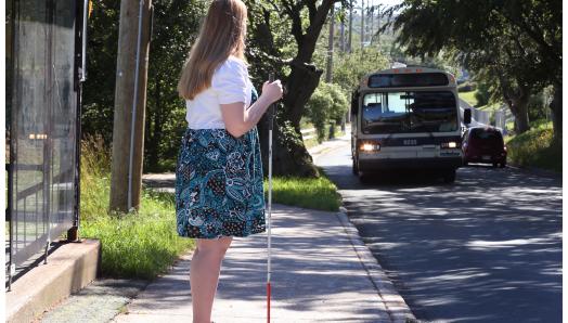 A woman holding a white cane standing at a bus stop waiting for the bus to pull up.