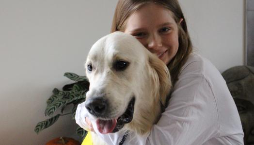Dani sitting and hugging her buddy dog George, a golden retriever wearing a bright yellow CNIB Buddy Dog vest, both smiling for the camera