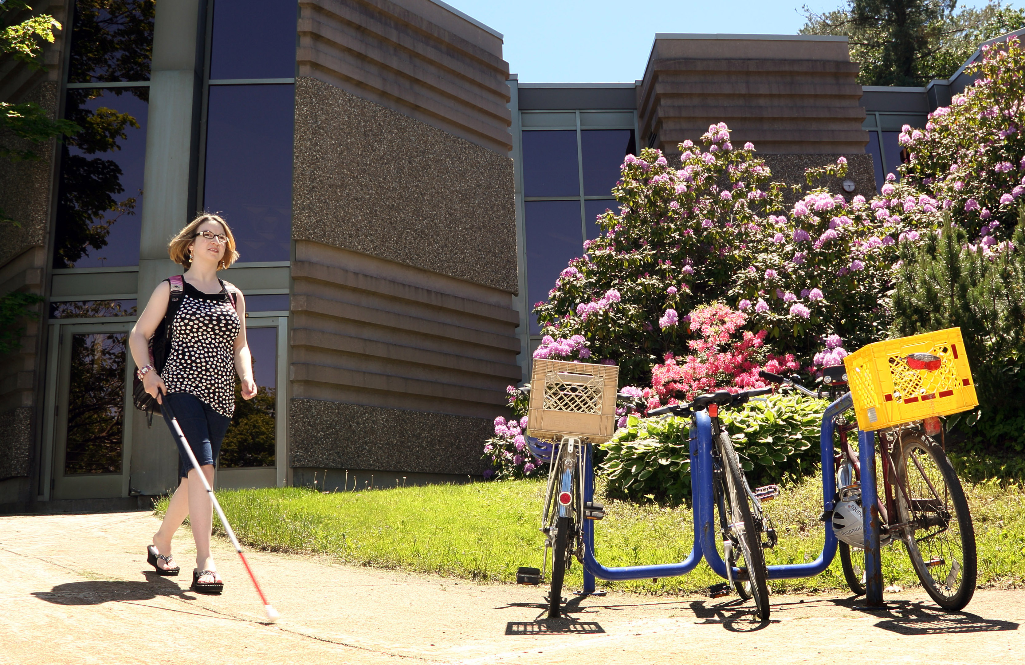 Young woman walks with white cane on campus while wearing a backpack.