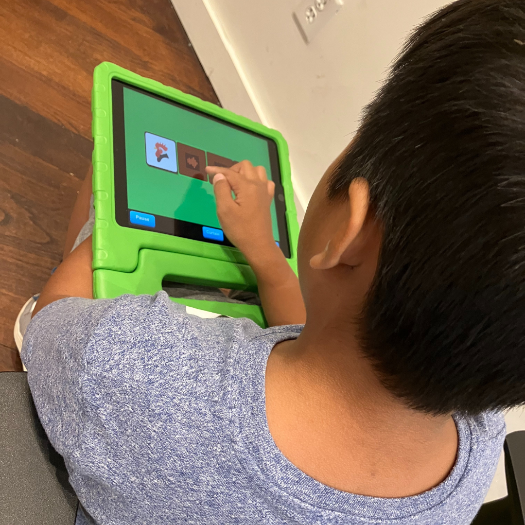 Un jeune garçon joue sur une tablette. Il est assis sur le sol, sa tablette posée sur les genoux est protégée par un étui en caoutchouc vert. Il tape sur l'écran pour interagir avec l'application éducative.