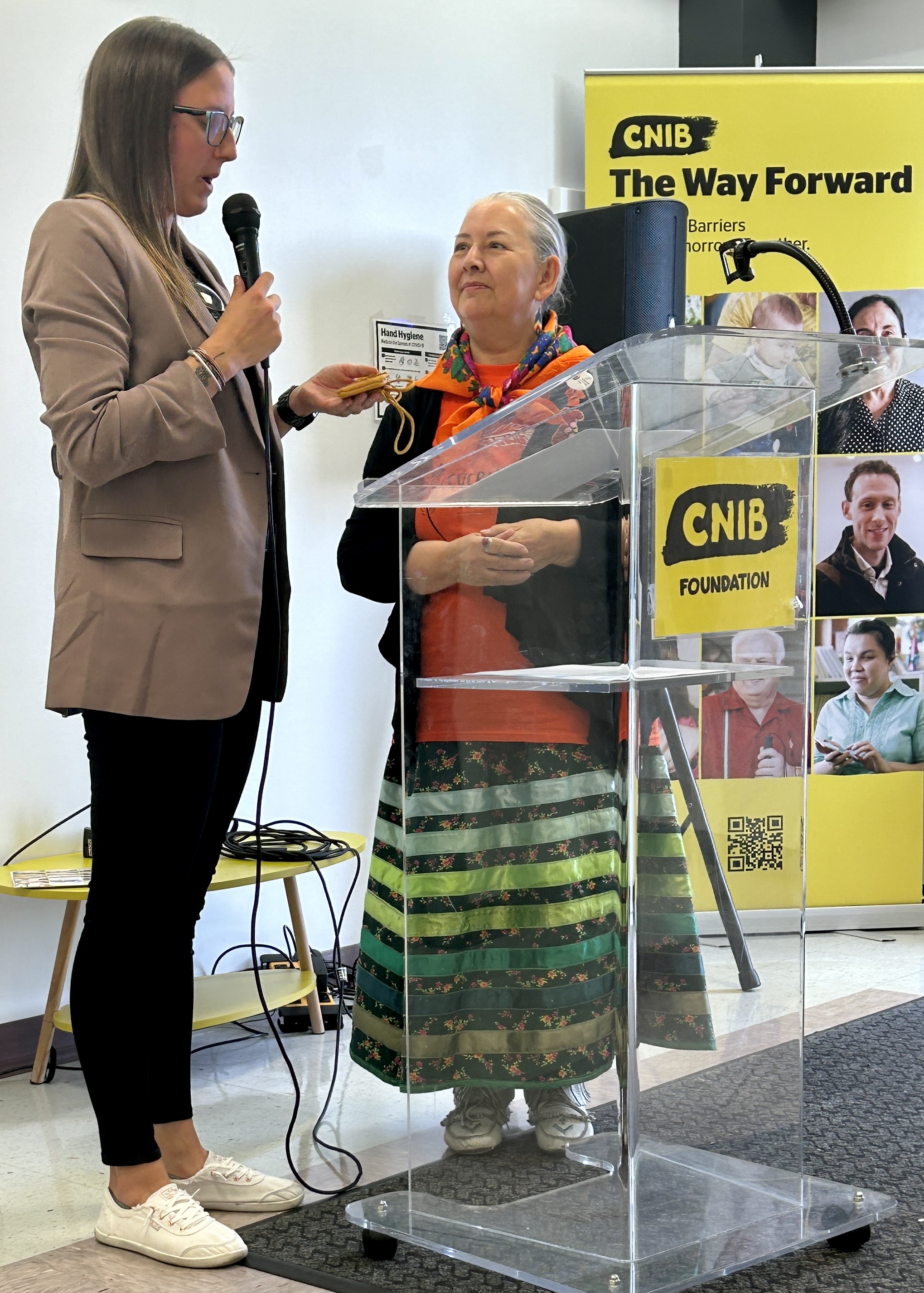 Une femme portant un blazer se tient à côté d'une aînée autochtone sur un podium avec une bannière d'INCA en arrière-plan.