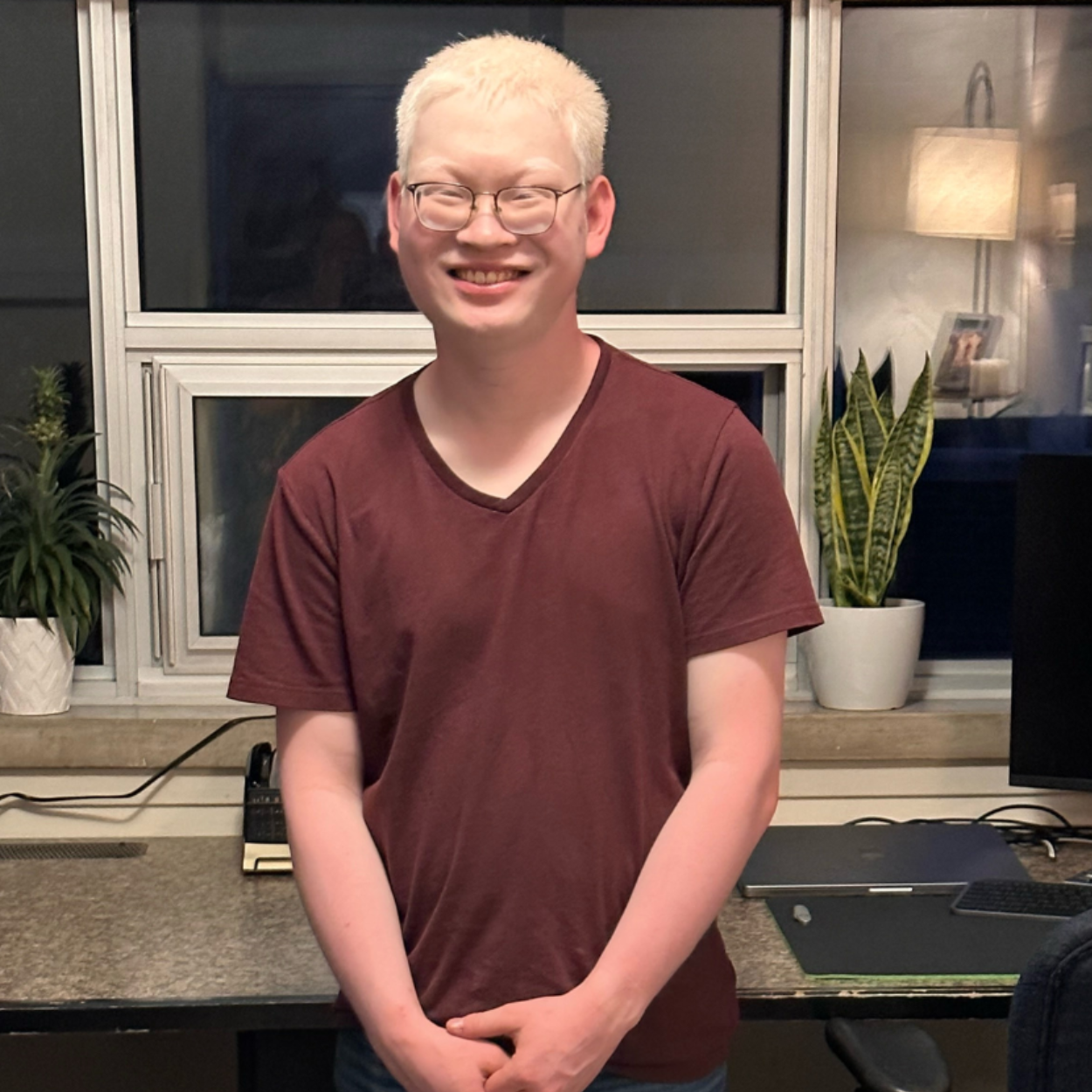 Adian stands in-front of a home office/computer workstation. He is smiling and standing with his hands clasped together. Aidan has platinum white hair, wears glasses and a brown t-shirt with jeans.