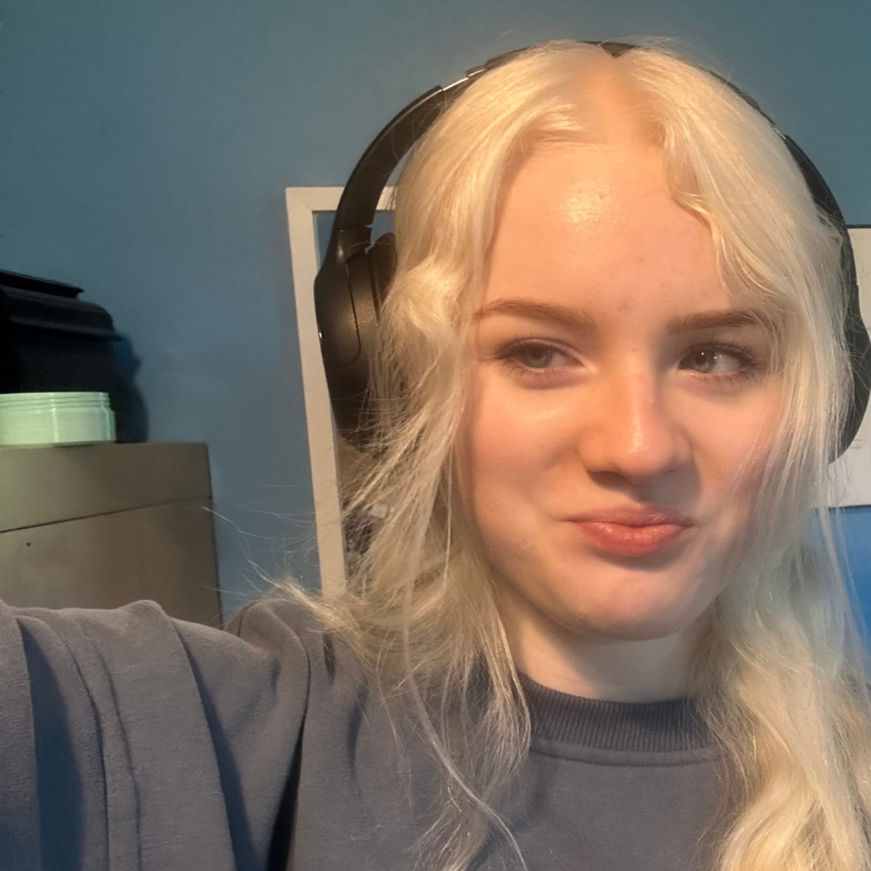 Acacia, une jeune femme aux cheveux blancs, sourit et pose pour une photo de type selfie. Elle porte des écouteurs sur les oreilles et un long sweat-shirt gris.
