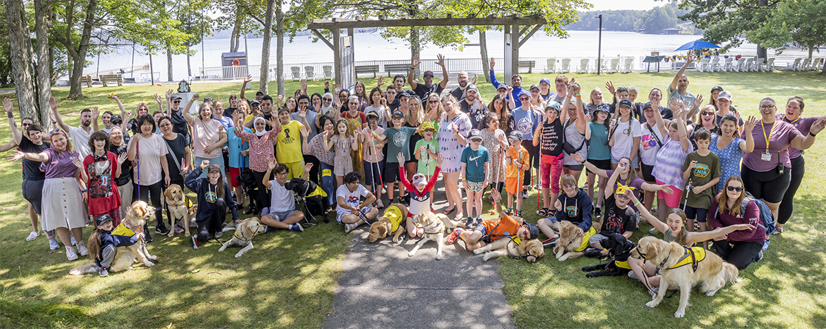Une photo de groupe réunissant les campeurs, le personnel et les chiens accompagnateurs, tous rassemblés sur la pelouse devant le bord de l'eau. Tout le monde sourit et lève les mains en l'air pour célébrer.
