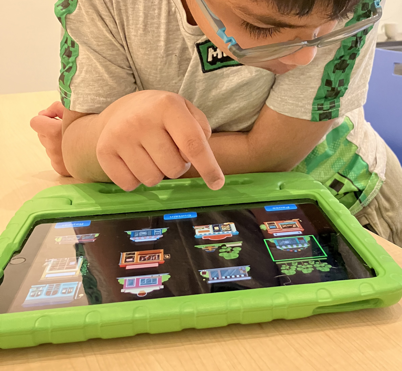 Un jeune garçon joue sur une tablette. La tablette est posée sur une table dans un étui de protection en caoutchouc vert. L'enfant tape sur l'écran de la tablette et interagit avec l'application Les 1ers taps.