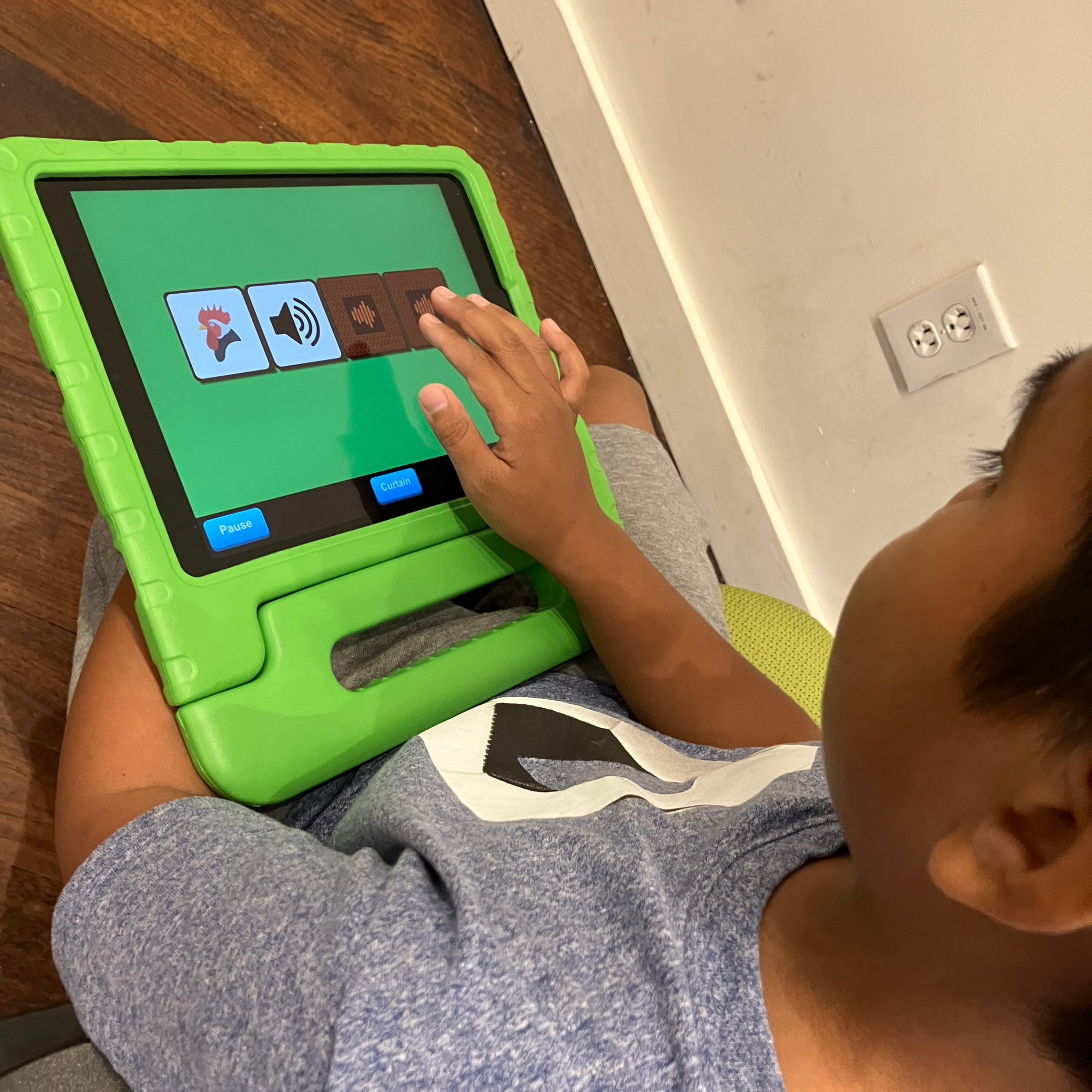 Un jeune garçon joue sur une tablette. Il est assis par terre et la tablette se trouve sur ses genoux, dans un étui de protection en caoutchouc vert. La photo est une vue aérienne de l'enfant qui tape sur l'écran de la tablette et interagit avec l'application Les 1ers taps.