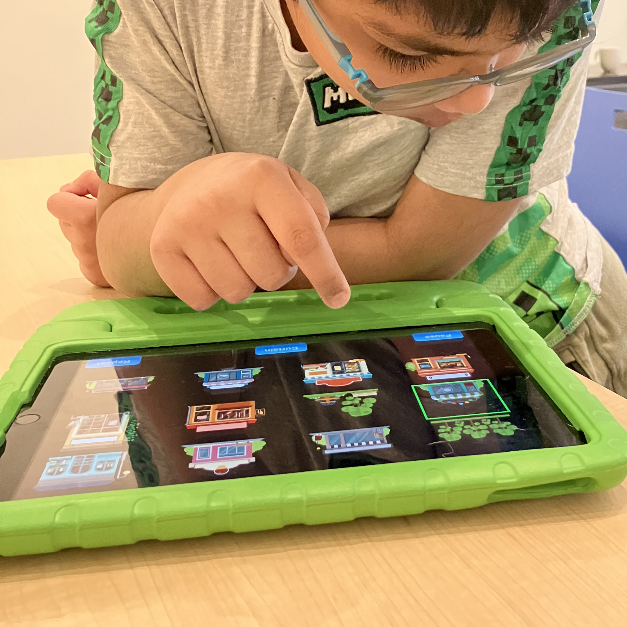 Un jeune garçon joue sur une tablette. La tablette est posée sur une table dans un étui de protection en caoutchouc vert. L'enfant tape sur l'écran de la tablette et interagit avec l'application Les 1ers taps.