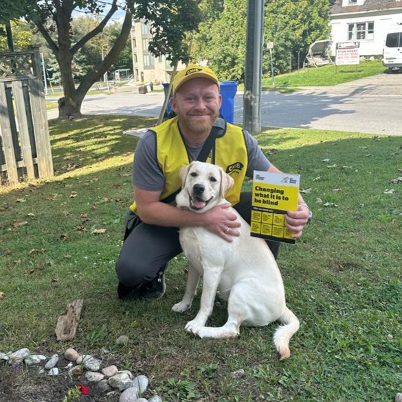 Un collecteur de fonds travaillant pour INCA est agenouillé à côté d'un chien sur une pelouse donnant sur la rue. Il porte une casquette et un gilet jaunes d’INCA et tient une grande carte présentant des faits sur la cécité.