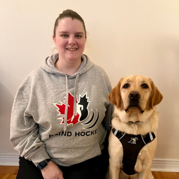 Laura et son chien-guide d'INCA, Toby. À l'intérieur, Laura est agenouillée sur le sol à côté de Toby, un labrador blond portant un harnais. Laura sourit et entoure Toby de son bras gauche. Laura porte également un chandail de Hockey sonore Canada. 