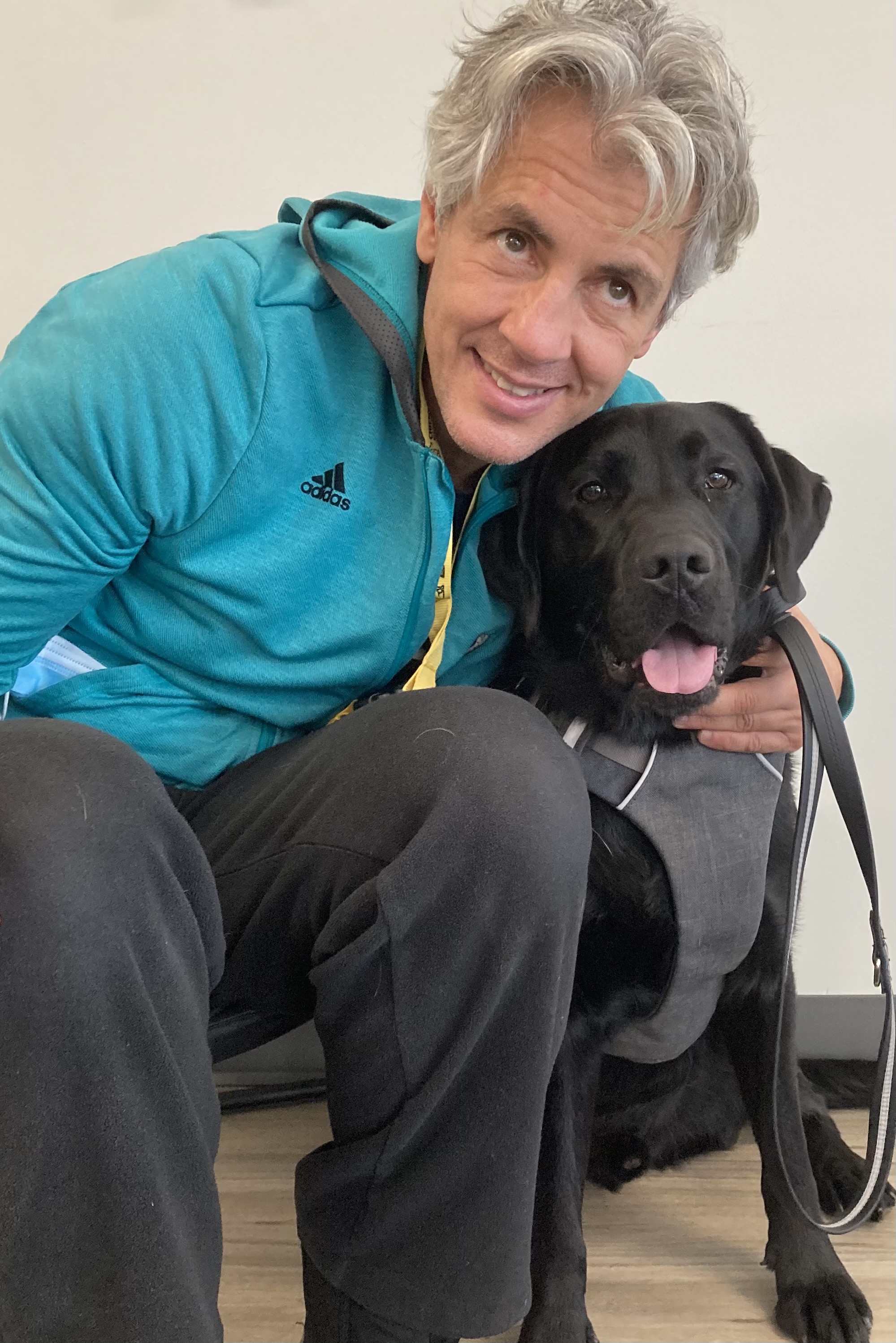 Une photo d’Ary, vêtu d’un veston Adidas turquoise et d’un pantalon noir, assis sur le sol, frottant le ventre de Don, un chien-guide d’INCA, et souriant pour la photo.