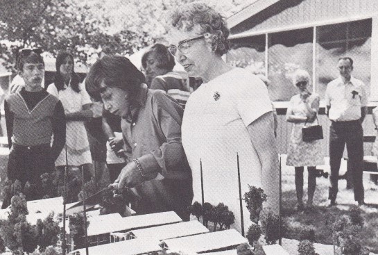A black & white photograph of Mary Patterson, Canadian Council of the Blind National President and William Dunlop, a young camper, cutting the special birthday cake – a realistic scale model of the Centre’s building baked for the occasion by CaterPlan Services/Division of CNIB.