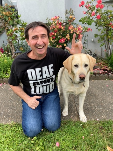 Eddy est assis devant un jardin dans une cour avec son chien-guide. Sa main gauche est au-dessus de la tête du chien et il signe « I love you » en langue des signes américaine. Le t-shirt noir qu’Eddy porte affiche le texte « Deaf and loud! » en gras. 