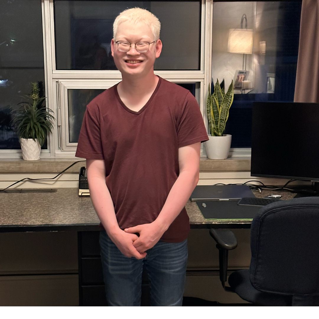 Aidan, un jeune homme aux cheveux blancs, se tient devant un bureau à domicile/un poste de travail informatique. Aidan porte des lunettes et un t-shirt marron avec un jean. Il sourit et se tient debout, les mains jointes.