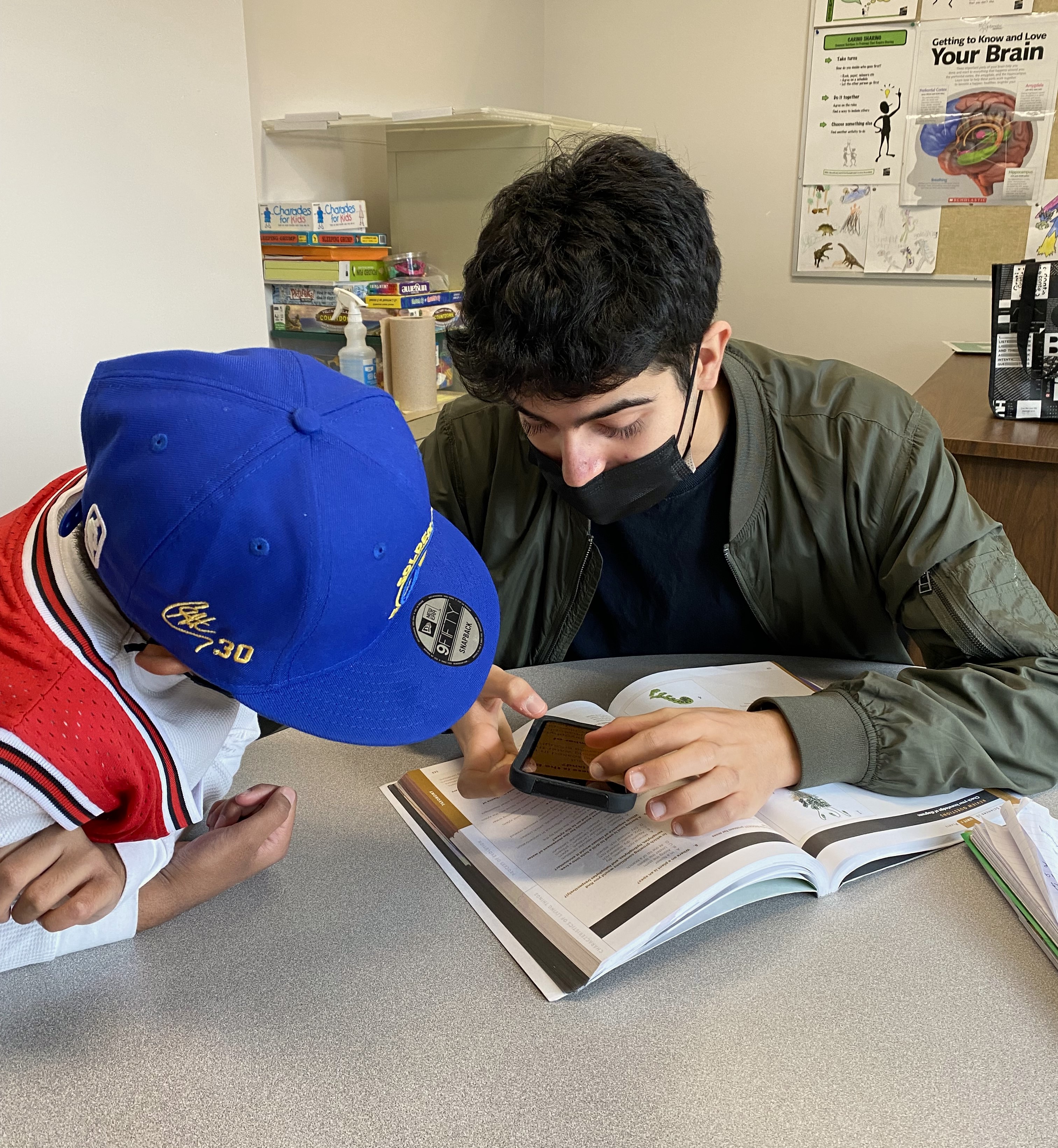 In a classroom, Aidan sits with a young student and shows him the magnification features on his iPhone. Aidan is holding his iPhone over a textbook.  The student intently leans-in for the demonstration.