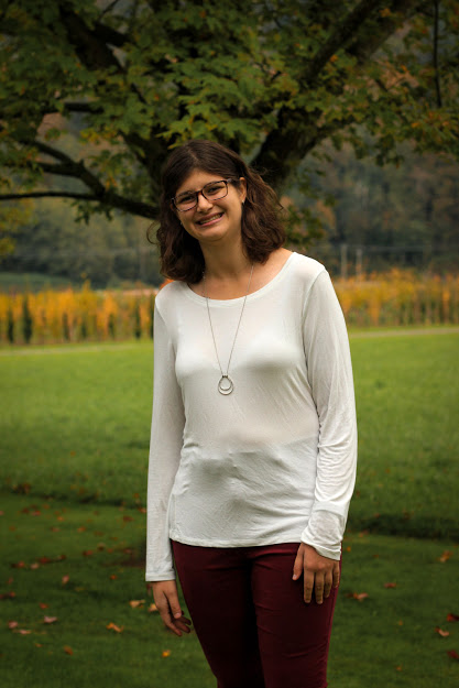 Amy Verwoerd poses for a photo outdoors with a lush green landscape of trees and grass in the background.