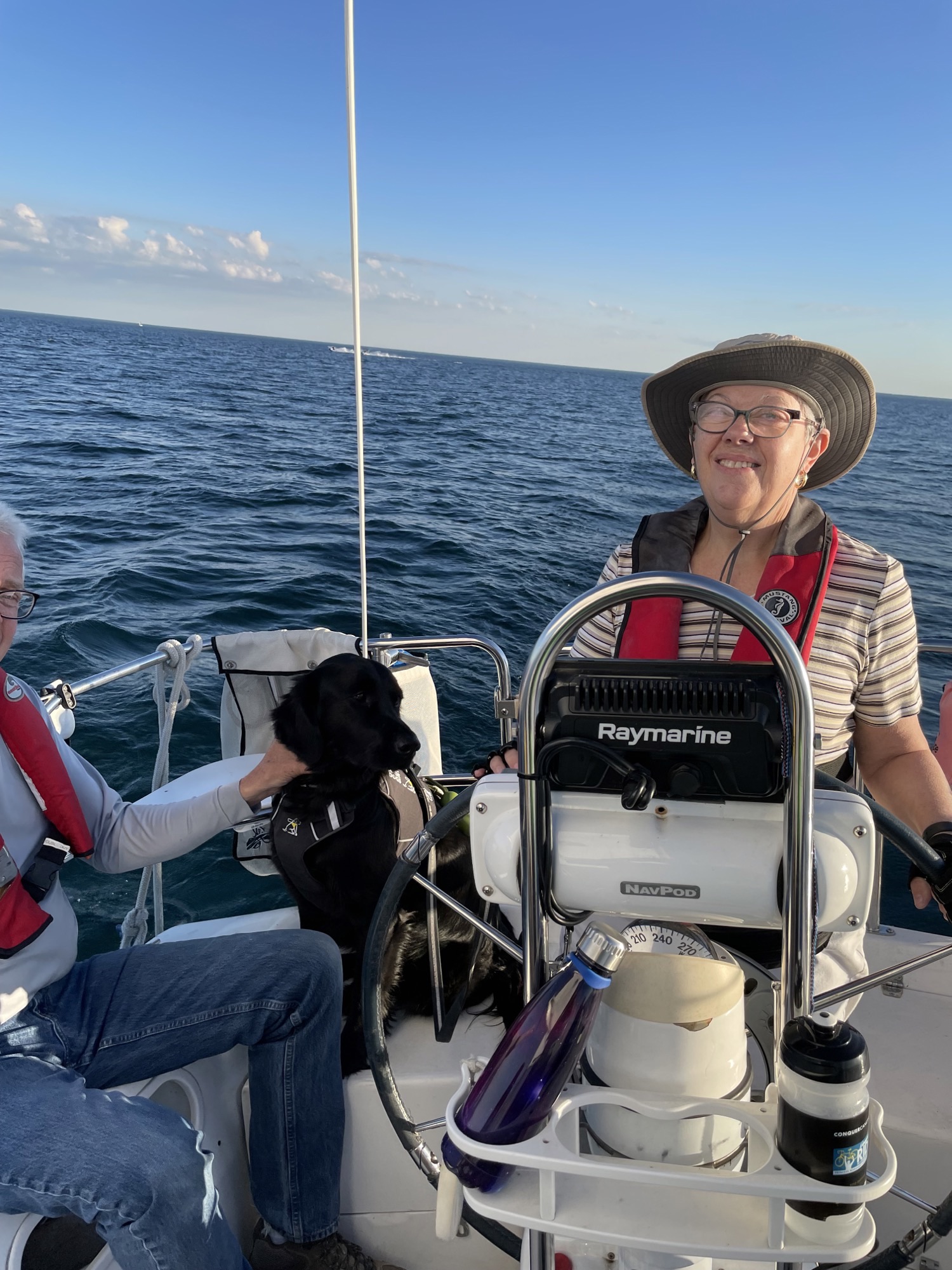 En naviguant sur le lac Ontario, Penny prend la barre du voilier. Elle porte des lunettes, un chapeau et un gilet de sauvetage rouge. Derrière Penny se trouve Honour, son chien-guide noir. À la gauche de Penny, une personne qui est visible en partie porte un gilet de sauvetage. C’est une journée ensoleillée, avec un ciel bleu clair, et le lac est calme.