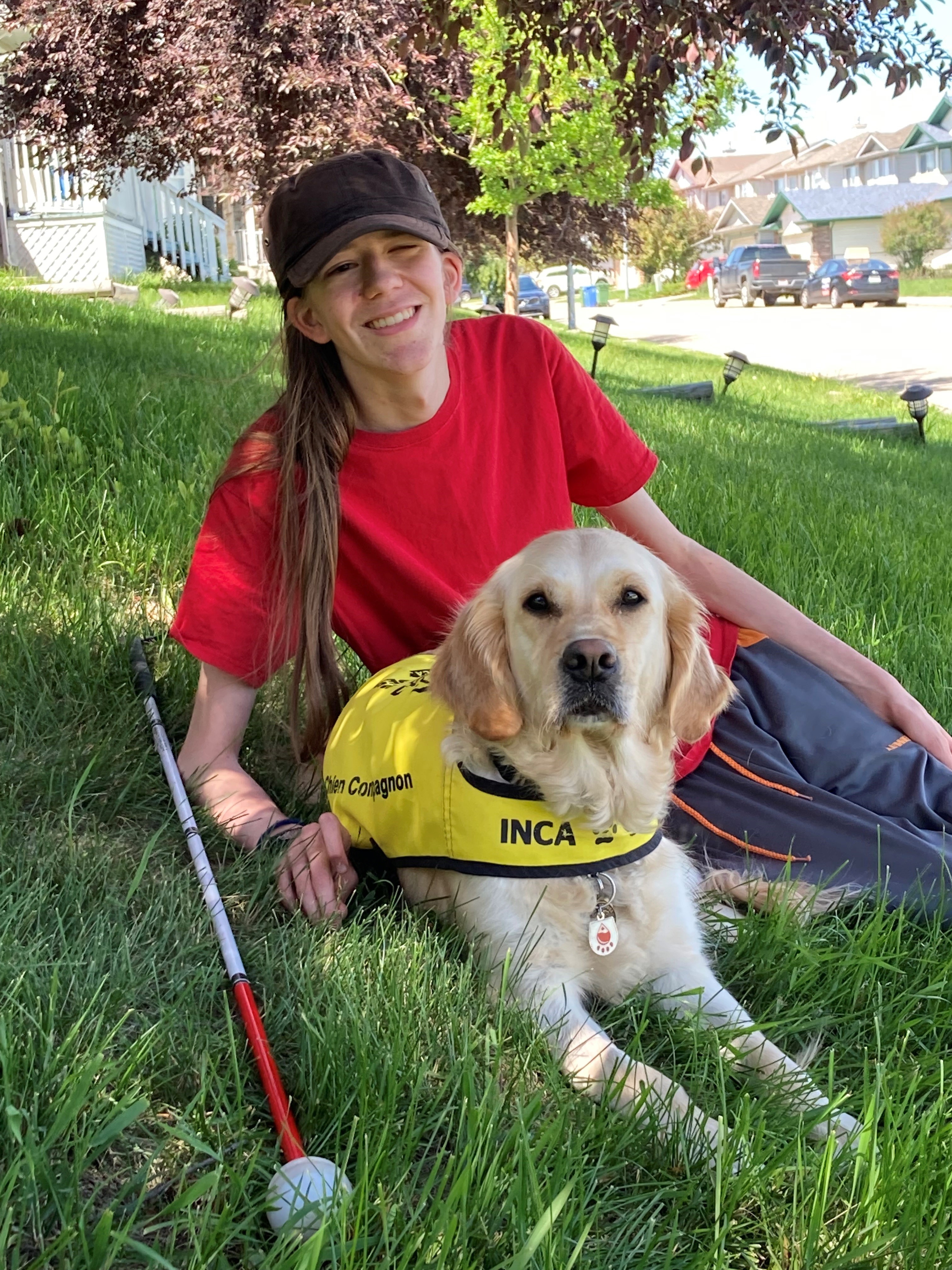 Zach est allongé sur l'herbe avec son chien Elsie, un golden retriever qui porte un gilet jaune d'INCA. La canne blanche de Zach est posée sur l'herbe près de lui.