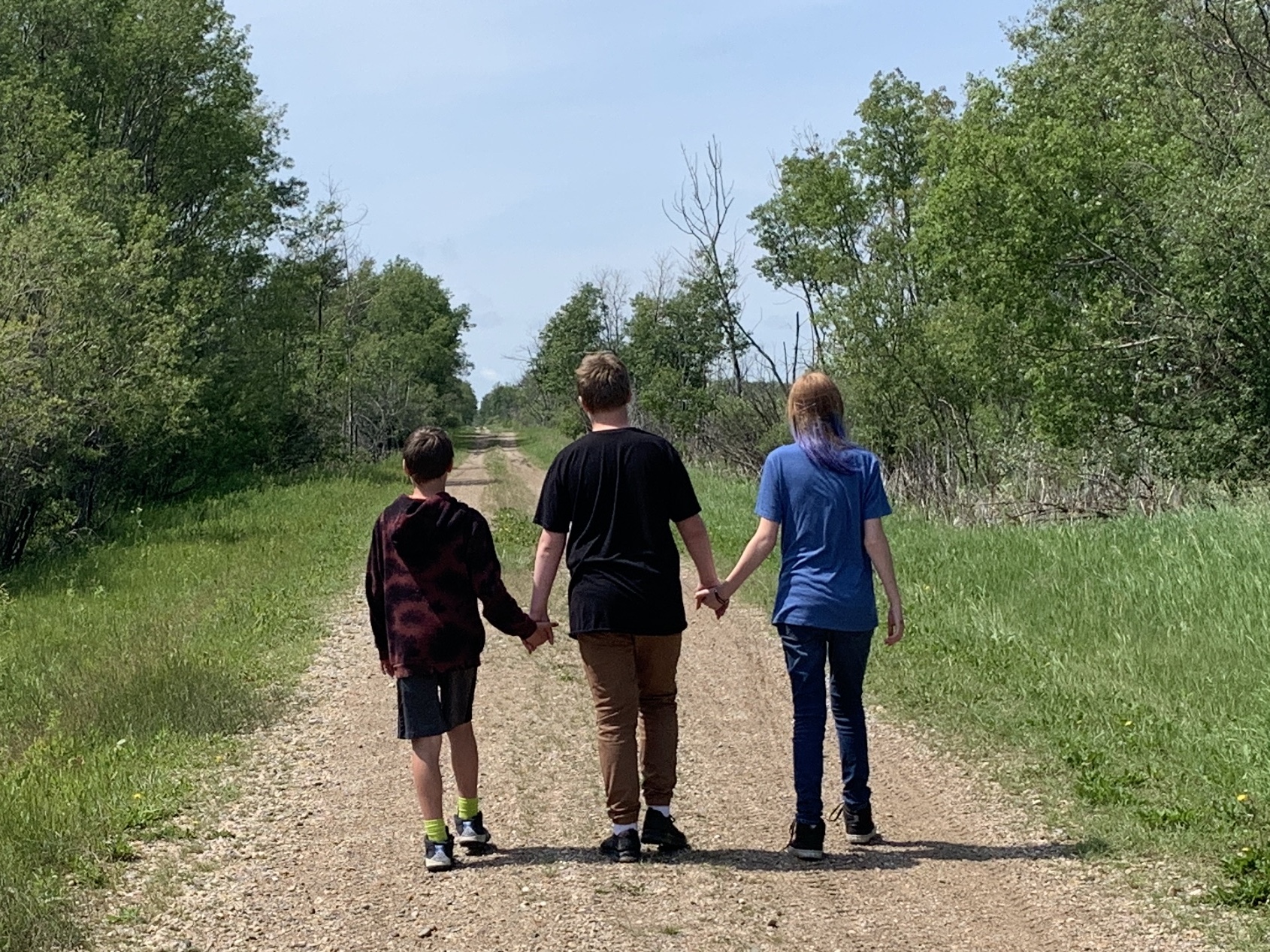 The backs of Ashley’s three children as they walk down a secluded road in a lush, green area. The children are holding hands.