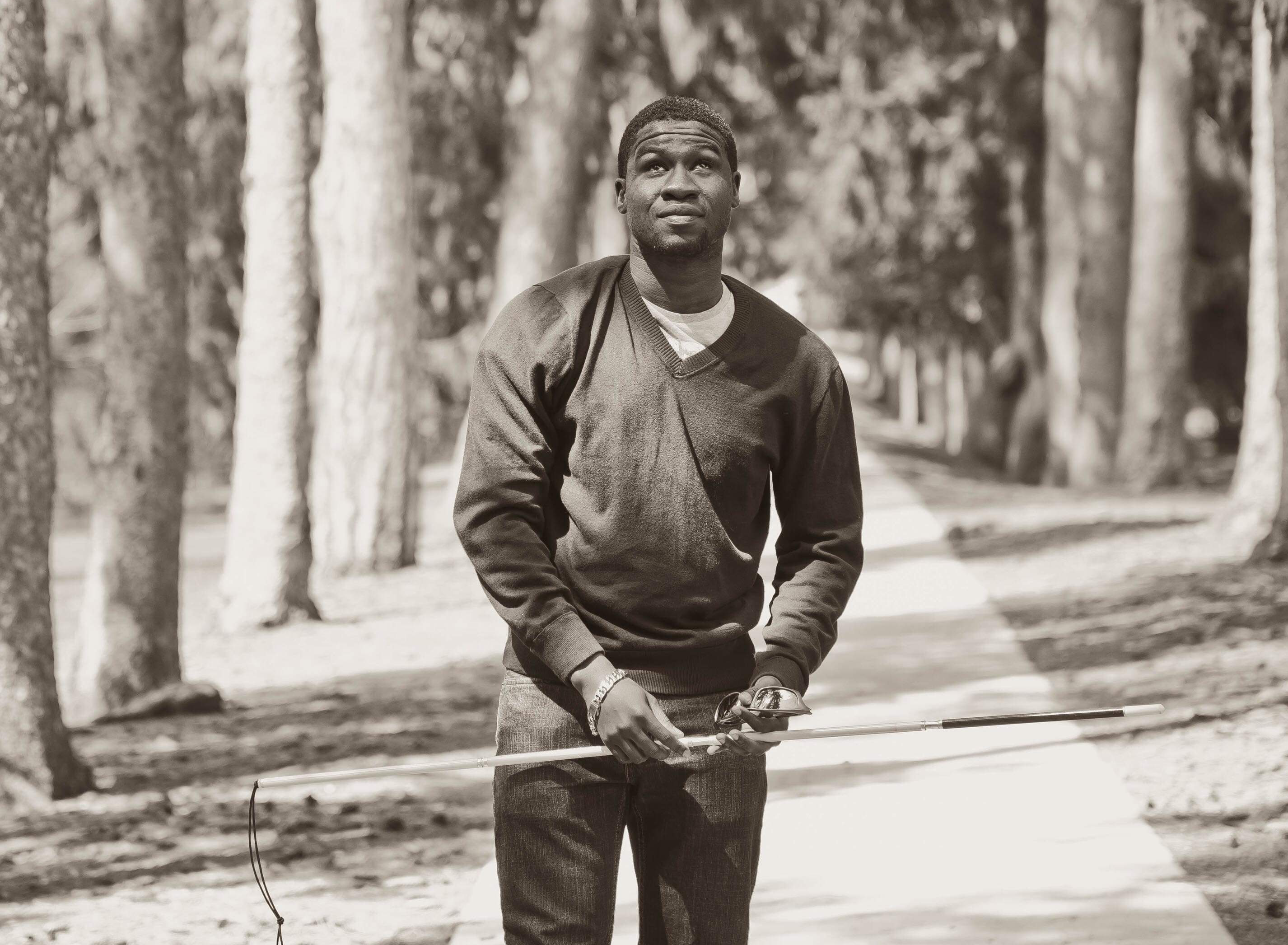 A black and white photograph of Ben walking in a forest. He holds his white cane up in the air near his waist.