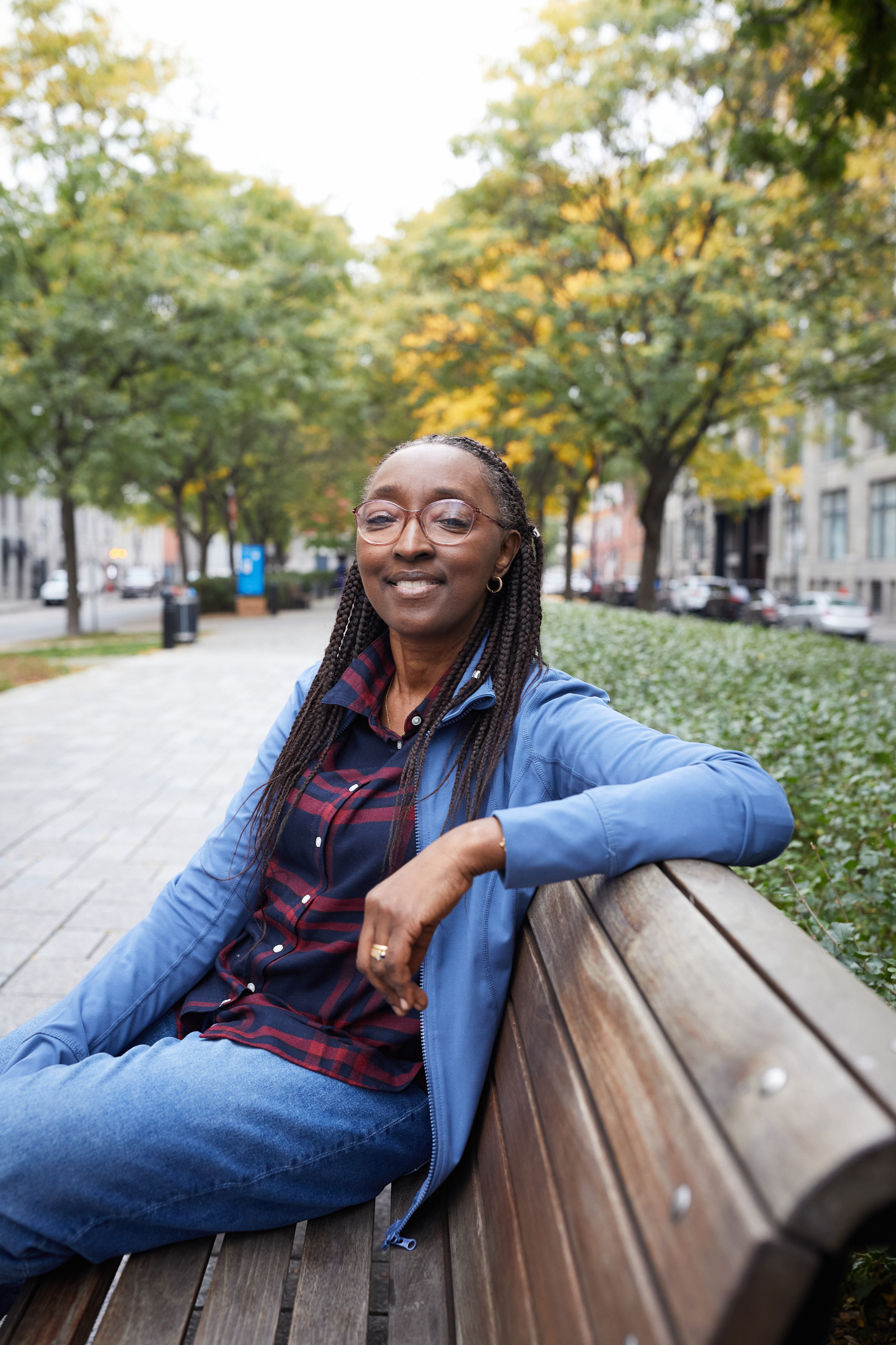 Binta est assise sur un banc extérieur en bois. Elle porte une chemise et des lunettes.