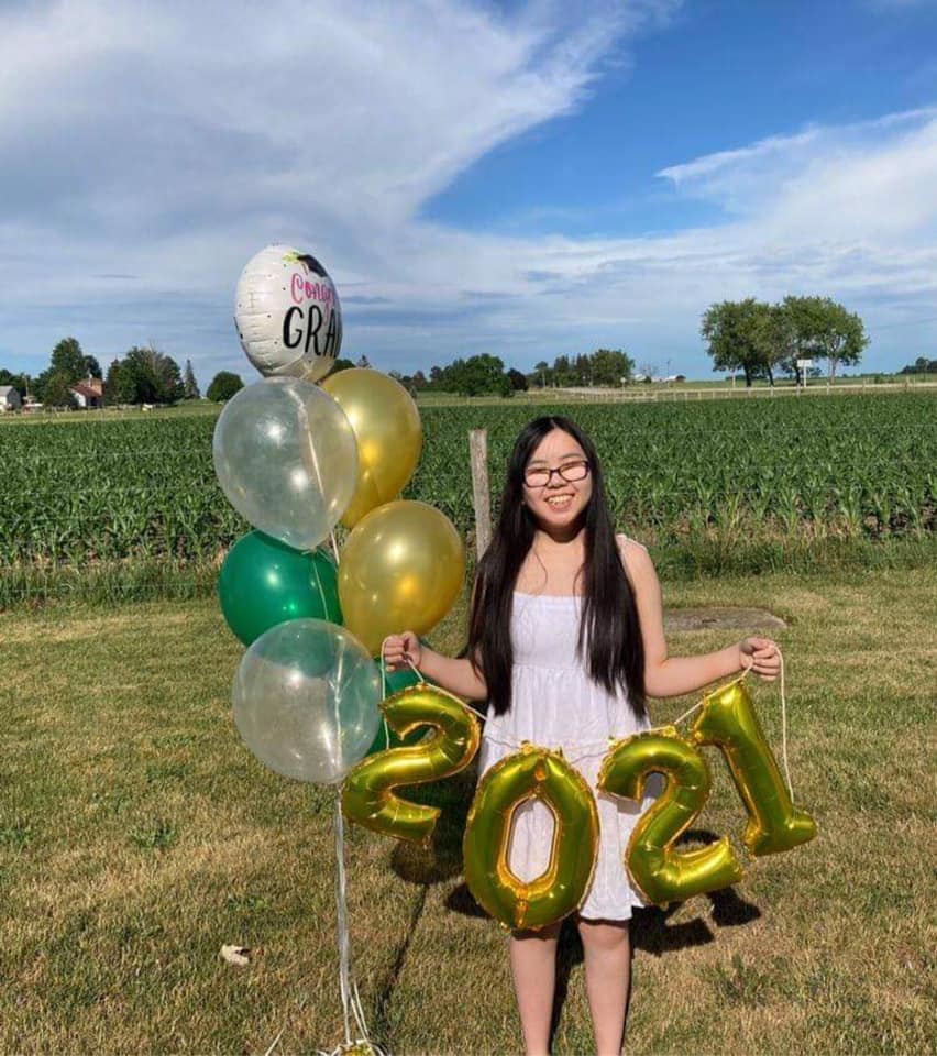 Stormy se tient devant un champ de maïs à la campagne par une journée d’été ensoleillée. Elle porte une robe                       bain-de-soleil et tient un bouquet de ballons sur le thème de la remise des diplômes.