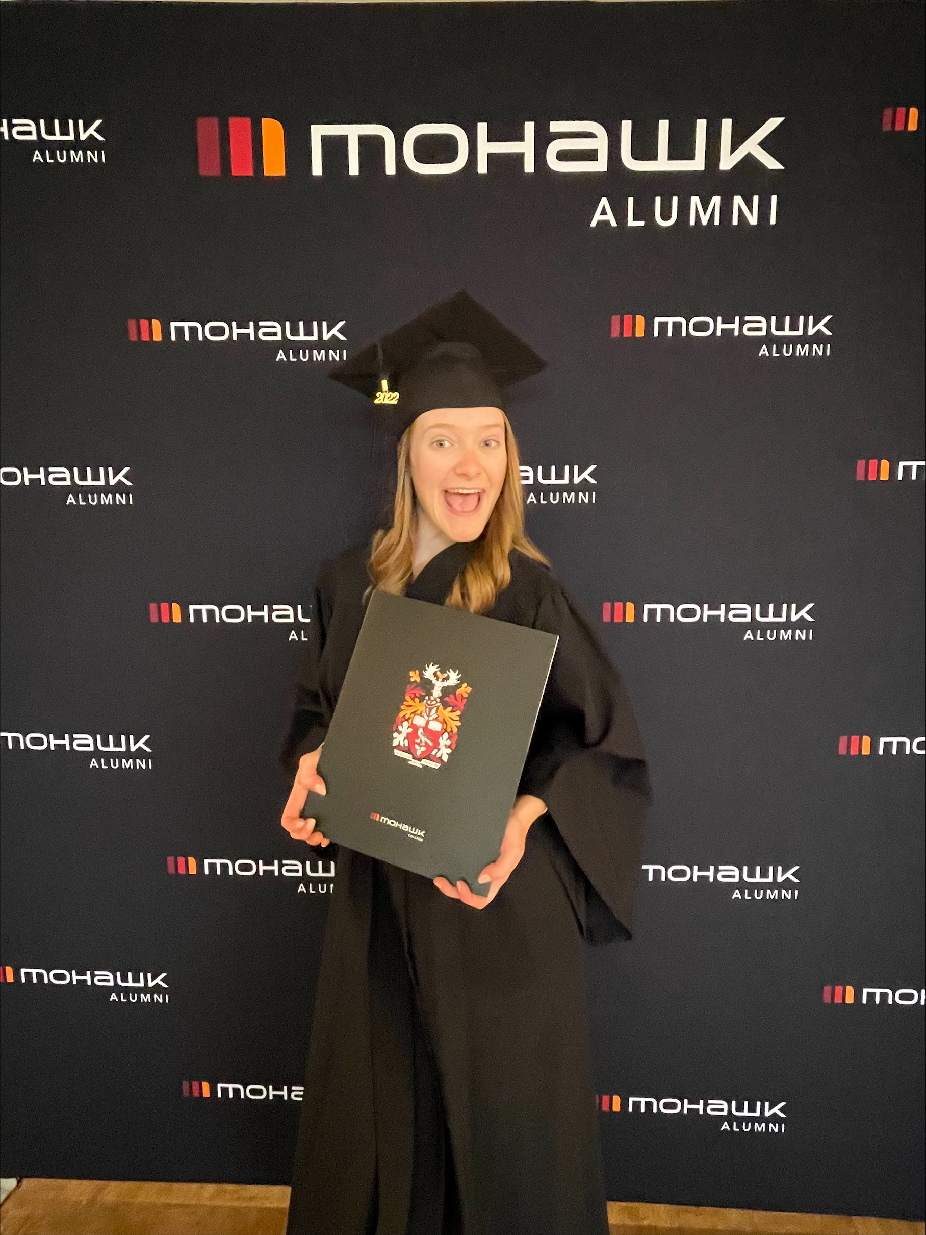 Jour de la remise des diplômes! Emilee, vêtue de son chapeau et de sa robe de diplômée, sourit et montre fièrement son diplôme. Elle se tient devant une toile de fond dotée du logo des anciens élèves du Collège Mohawk.