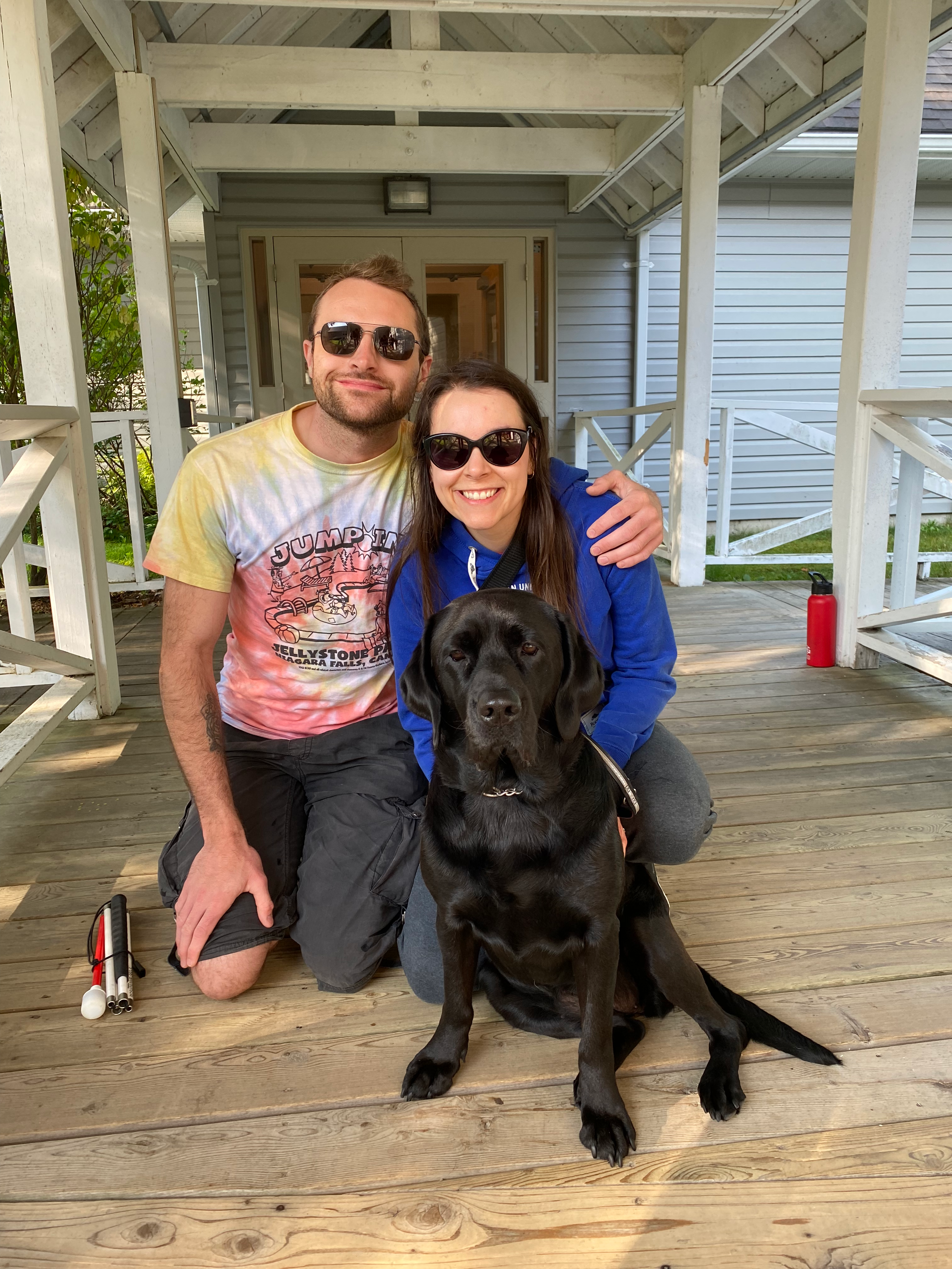 At CNIB Lake Joe, Micheal and Danica kneel on the boardwalk with a black Labrador Retriever. Picture taken during the Orientation & Mobility/Guide Dog Immersion program at CNIB Lake Joe.