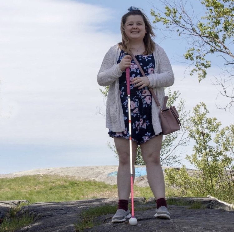 Alicia Chenier smiles and poses for a photo outdoors. Lush green trees and a blue sky in the background. She holds her white cane in her right hand.