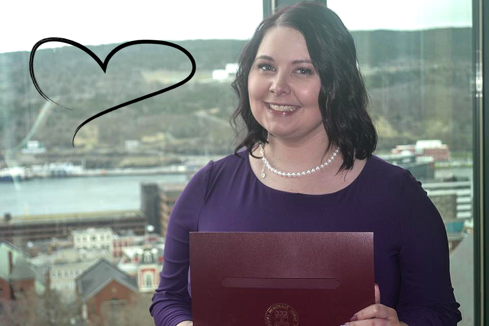 Brittany Farrell, souriant pour la caméra, debout devant une fenêtre qui donne sur la ville de St. John's (Terre-Neuve). Une image en forme de cœur est affichée dans la fenêtre