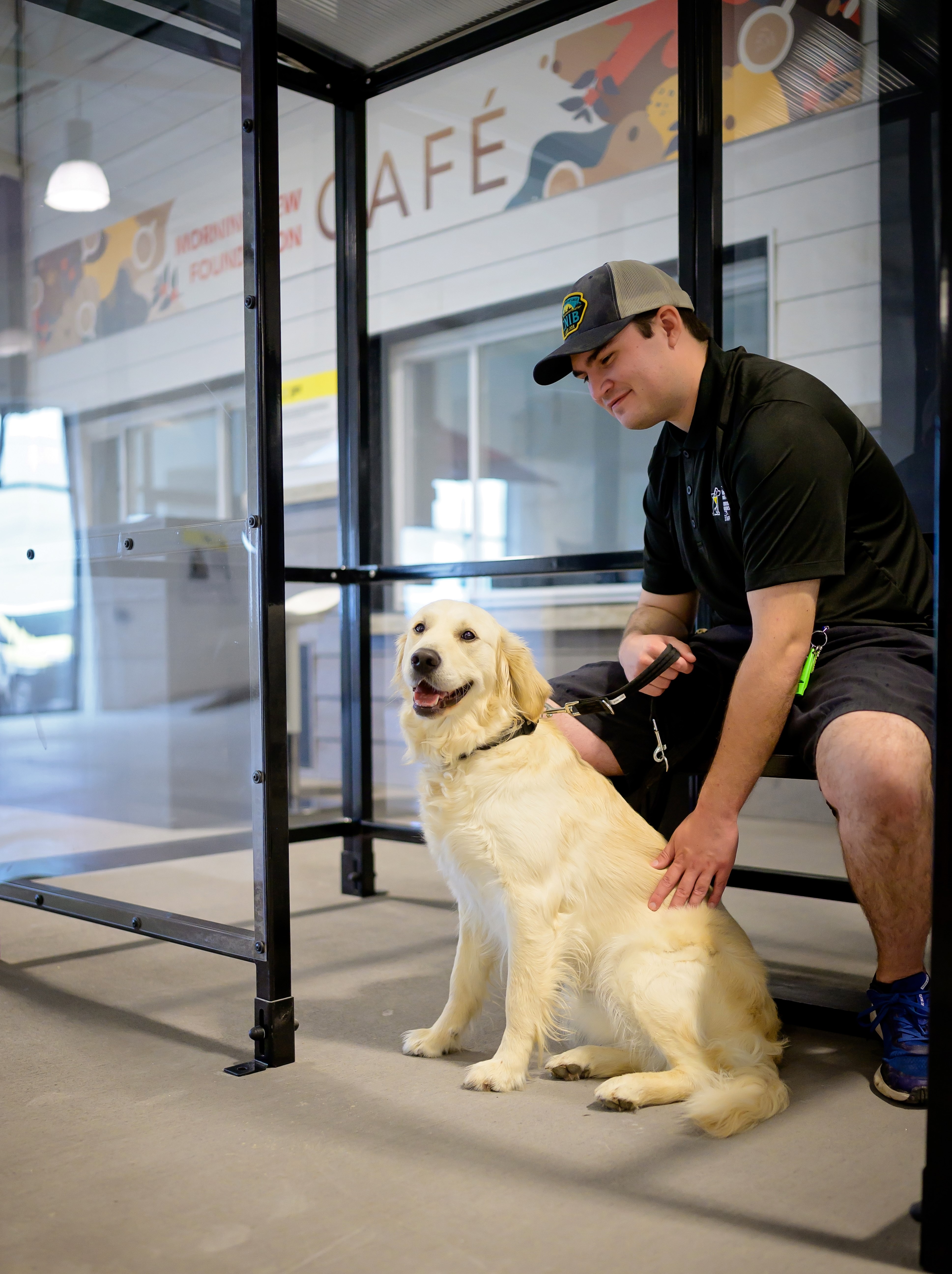  Un golden retriever souriant est assis à côté de son dresseur, qui porte une casquette et une chemise noire, à un arrêt de bus simulé à la ville intérieure des Chiens-Guides d’INCA. Une enseigne indiquant « Café » est visible à l'arrière-plan.