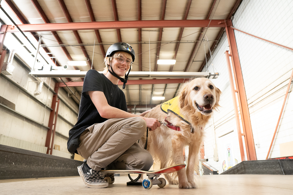 Un jeune homme à la peau claire, portant un casque noir et des lunettes, sourit et s'agenouille sur un skateboard à côté de son chien compagnon, un golden retriever. Ils se trouvent dans un skate park intérieur et le chien porte un gilet jaune de l’INCA.