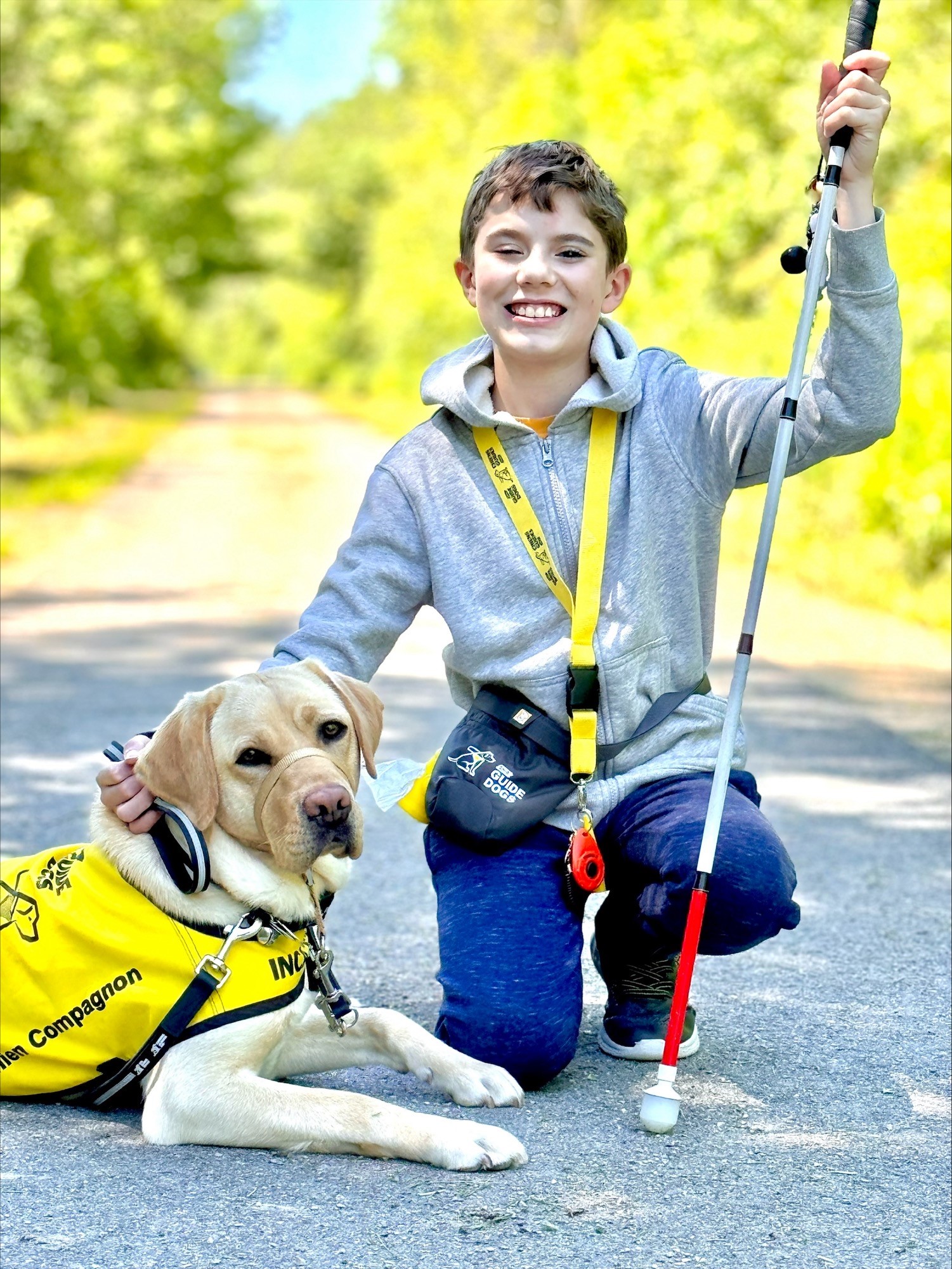 Caleb, un garçon souriant à la peau claire, tient sa canne blanche et s'accroupit à côté d'Amber, un labrador blond. Amber, allongée, regarde la caméra avec une expression sérieuse tout en portant son gilet jaune et noir de chien compagnon d'INCA. Ils posent sur une route pavée tranquille avec des arbres verts derrière eux.