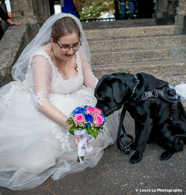 A photo of Christine on her wedding day, kneeling next to Edie in her wedding dress holding a beautiful bouquet of pink and blue flowers.