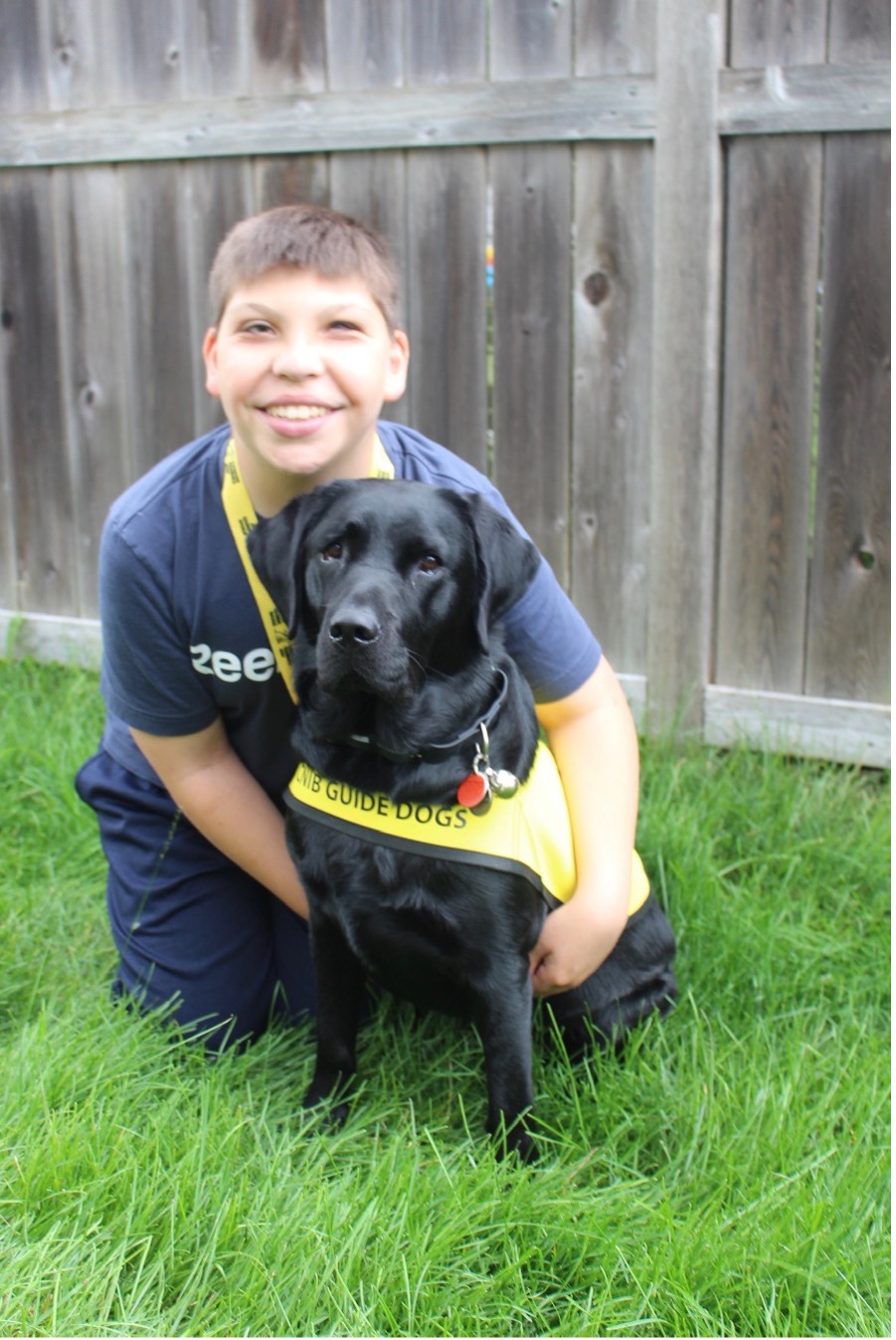 Connor is looking at the camera and has his arms around June, giving her a big hug; June, a black lab, is sitting looking at the camera.