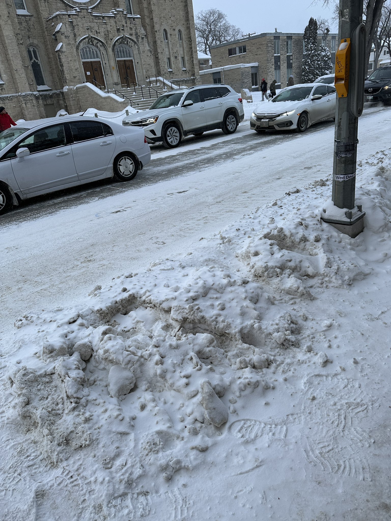 A snowbank along a sidewalk in Winnipeg. The snowbank obstructs both the path of travel and a pole that houses the pedestrian crossing push-button.