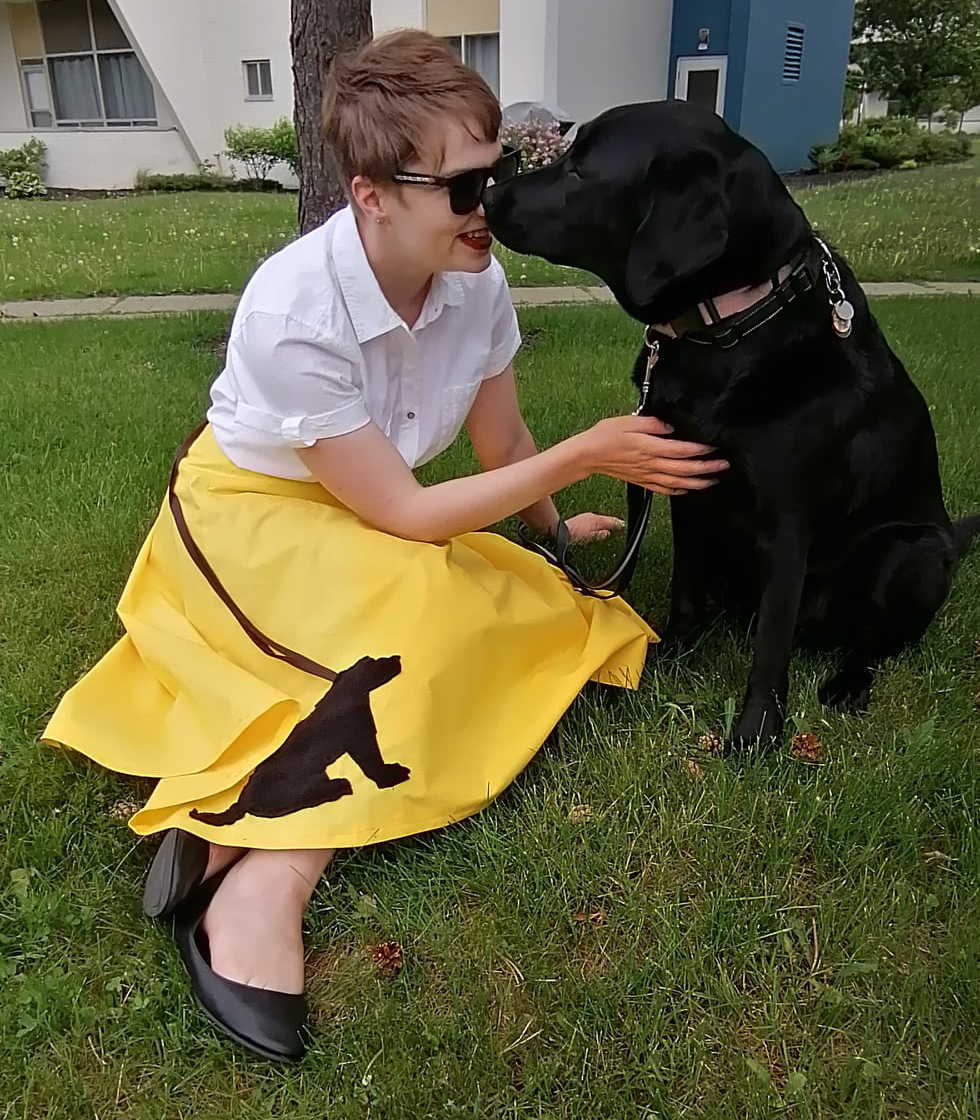 Denise, assise dans l'herbe, vêtue d'une jupe jaune vif, nez-à-nez avec Tara, son chien-guide d’INCA. 