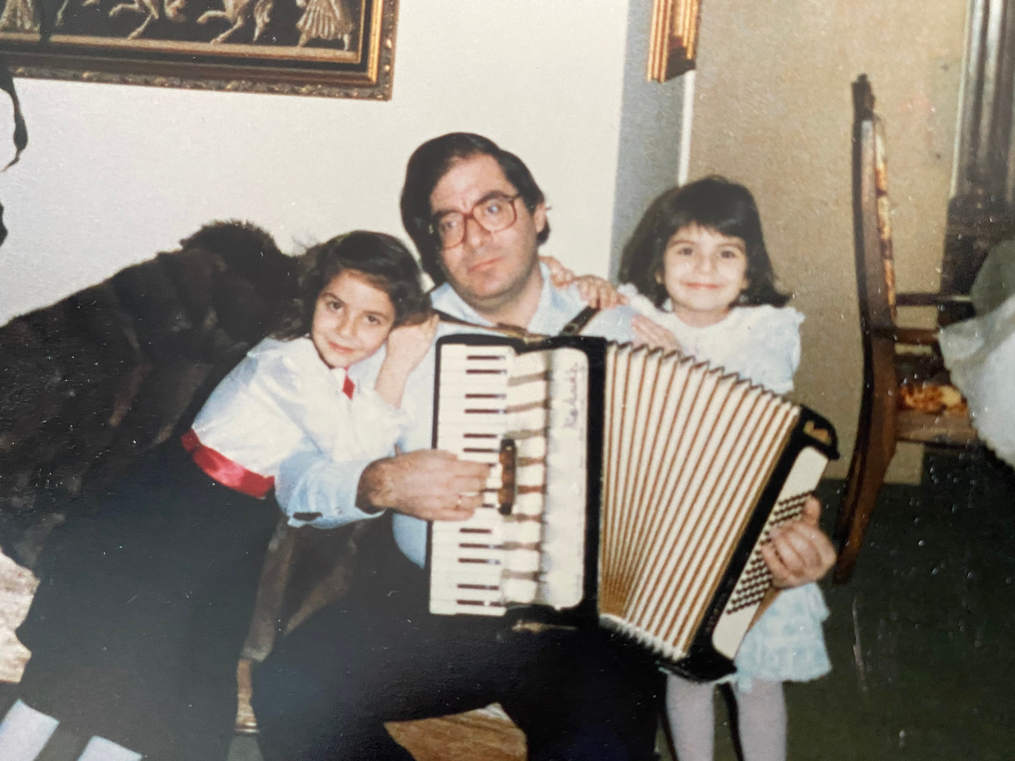 A photograph from the late ‘80s. Dimitrios plays the accordion. A young Angela sits on the left. Her sister sits on the right. 