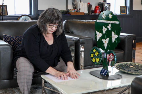 Robyn est assise sur une chaise revêtue de cuir et lit la description en braille d’une sculpture tactile que le canton d’Oro-Medonte lui a demandé de réaliser. La sculpture, posée sur la table, comporte des fleurs tactiles : des trilles, des boutons d’or et un cypripède.