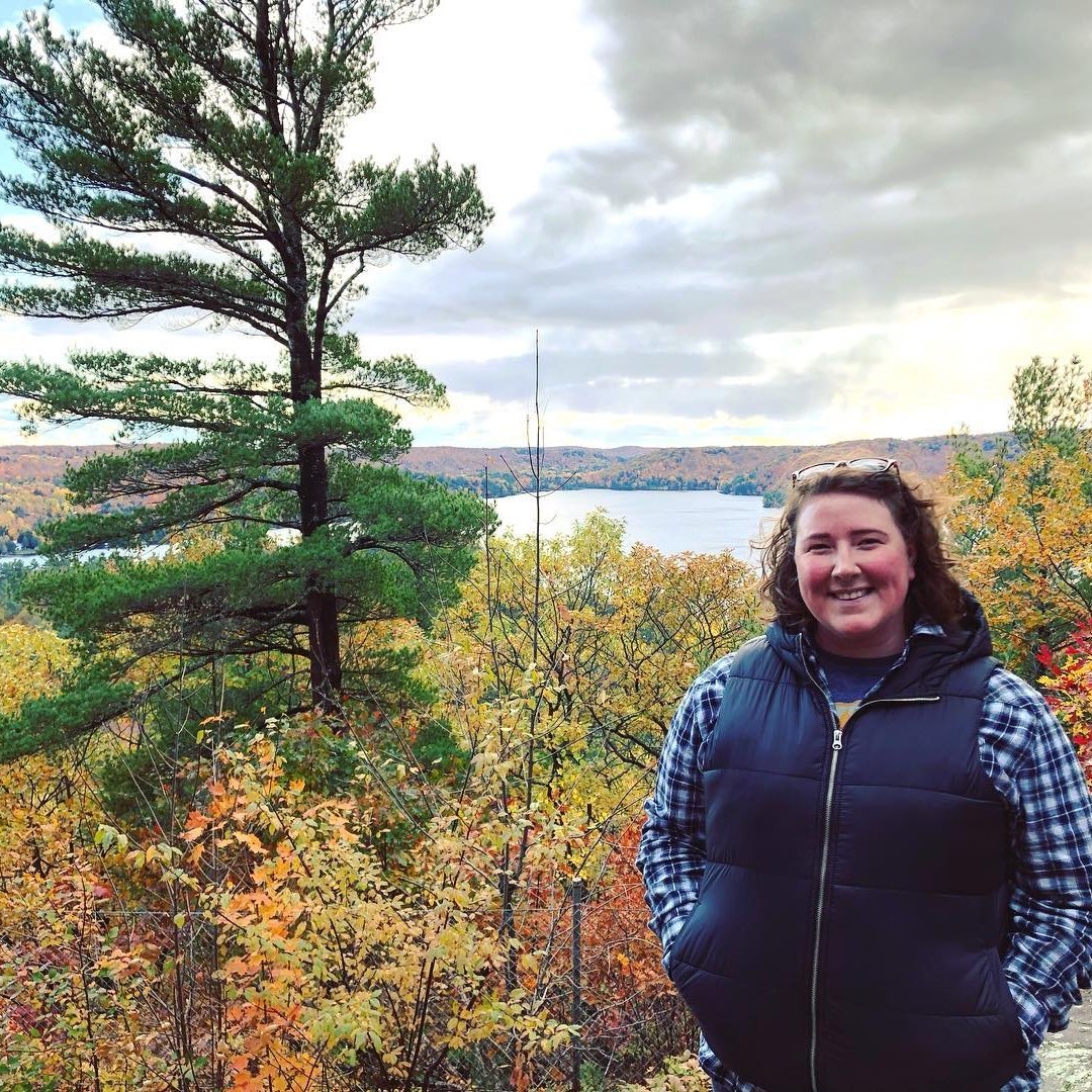 Emily on a hike in the fall. She poses in front of a lush area of trees. A lake is in the background.