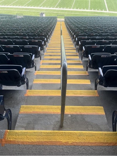 Une volée d’escaliers de stade en béton, dont le sommet donne sur un terrain de football, avec des rangées de sièges en gradins de part et d’autre. Les escaliers sont séparés au milieu par une rampe et chaque escalier est peint d’un jaune doré brillant sur toute la longueur des bords avant.