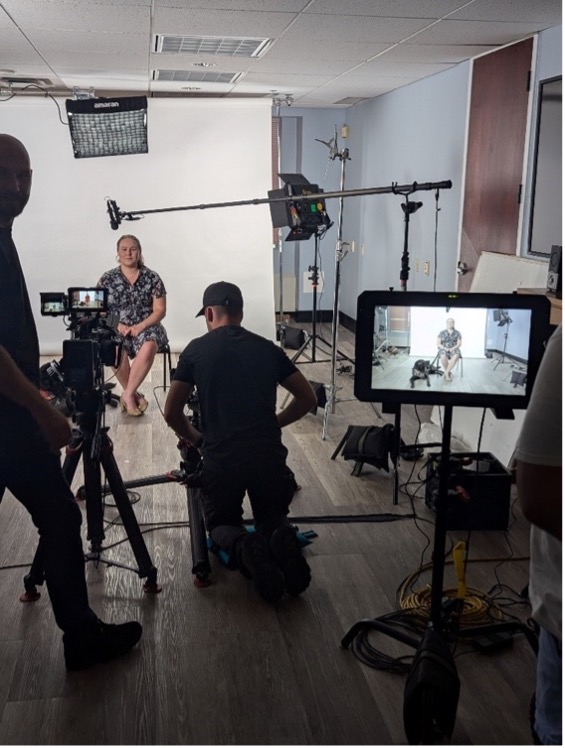 Taylor Bauer et son chien-guide Wallace sont interviewés aux fins d’une vidéo d’éducation publique. Taylor est assise avec un écran blanc derrière elle, tandis que plusieurs membres du personnel de production font fonctionner une caméra et du matériel d’enregistrement devant elle.