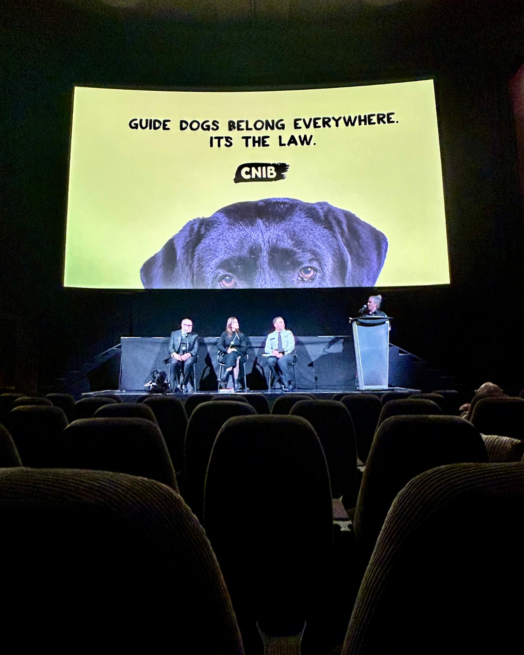 Trois personnes sont assises sur une scène en attente de participer à une table ronde. Un modérateur se tient à leurs côtés, derrière un podium. Une photo de la tête d’un chien-guide avec les mots « Guide dogs belong everywhere. It’s the law » (Les chiens-guides sont à leur place partout. C’est la loi) est projetée derrière eux.