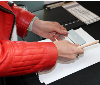 Une femme tient un guide de signature et un gros crayon par-dessus une page de papier. À ses côtés se trouve un bulletin de vote en gros caractères et un gabarit de vote tactile.
