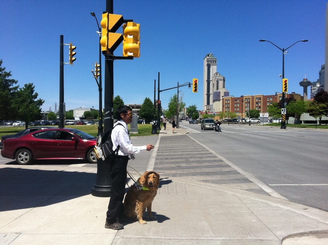 Un jeune homme et son chien-guide au coin d’une intersection très fréquentée. Dans sa main droite, il tient un porte-clés Key2Access. Dans sa main gauche, il tient le harnais de son chien-guide