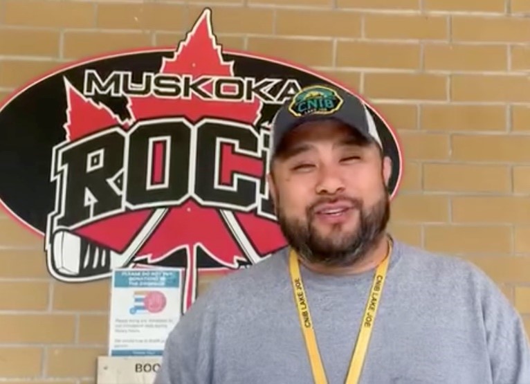 Eugène, qui porte une casquette de baseball, un chandail et un cordon de l'INCA Lake Joe, se tient à l'extérieur d'un bâtiment derrière lequel se trouve le logo " Muskoka Rocks " (un cercle noir avec une feuille d'érable rouge et deux bâtons de hockey).