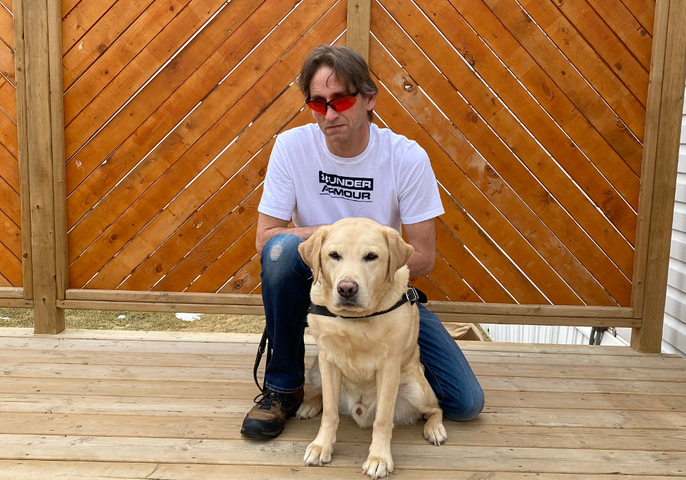 Gary, debout à l'intérieur contre un mur de briques avec son chien-guide. 